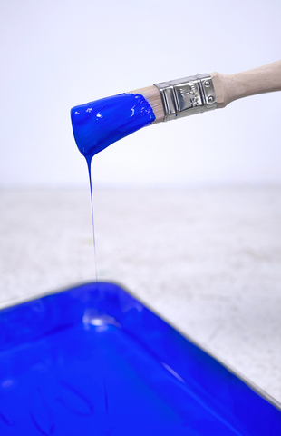 A close up of a small paint brush saturated with ultramarine blue paint, dripping onto a paint tray filled with electric blue paint
