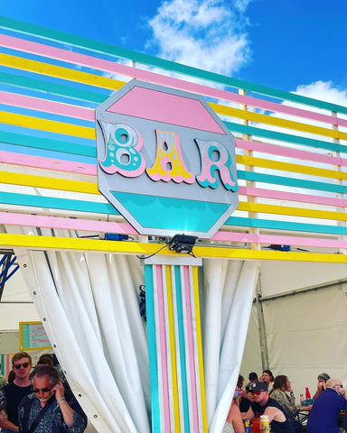 An image of an outdoor bar space with colourful thin wooden strips painted in pink, yellow and aqua colours, a circle sign with circus-type font reading 'bar' and white curtains, with a large group of people photographed in the background