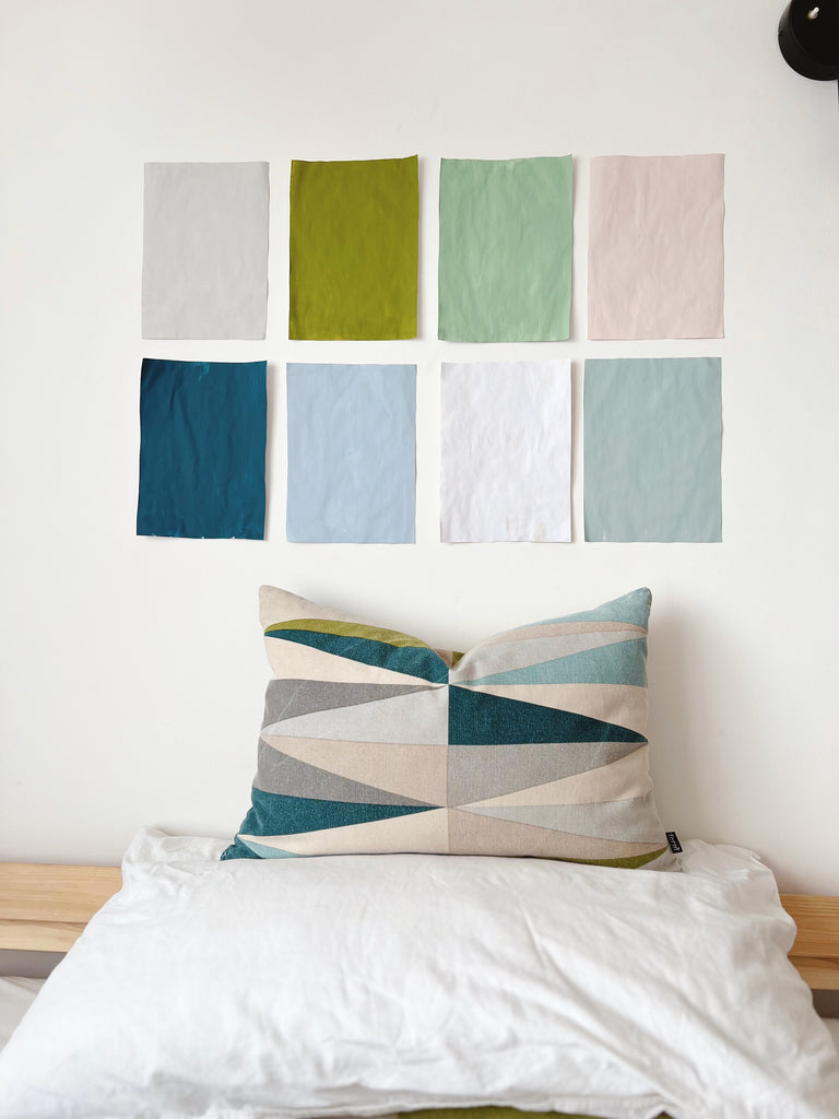 four coloured rectangular paper sheets painted with YesColours neutral, green, blue and pink paint colours, taped onto a white wall above a bed with geometric pattern pillow placed on top of a white bedding