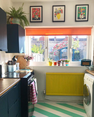 photo of a small kitchen space decorated with different bright colours - dark grey cupboards, wooden worktops, electric yellow radiator on the neutral wall, bright orange blinds and sun reflecting on the stripes on the floor, painted in white and mint green