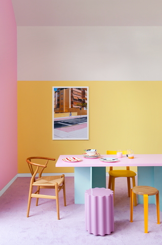 Multicoloured living room with a pastel pink wall on the left and a split-colour central wall with top half white and bottom half yellow; decorated with a long rectangular table with pink tabletop and aqua coloured legs, vintage wooden chairs, lilac geometric shaped stools and pink carpet