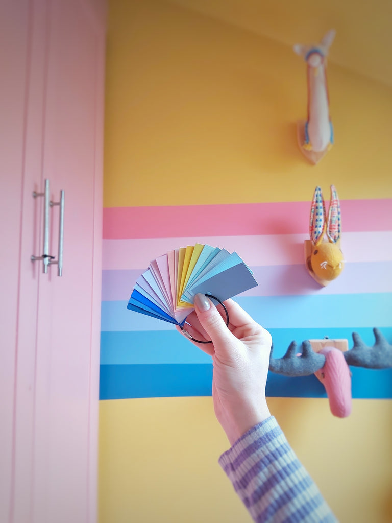 female hand holding a selection of paint colour chips against a yellow wall in a nursery room with a painted rainbow on it