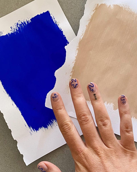 Woman's hand photographed above two pieces of lining paper - one painted in royal Electric Blue paint and the other one painted in warm, dark neutral colour