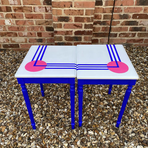 upcycled nest of mid-century tables in white, ultramarine blue and hot pink colours, arranged together on a pebble surfaced and in front of a brick wall