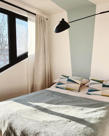 photo of a small bedroom with grey-green bedding, geometric pattern in teal on neutral pillowcases, a muted green geometric line painted on a neutral wall behind the bed and a black window frame looking out at the natural sunlight