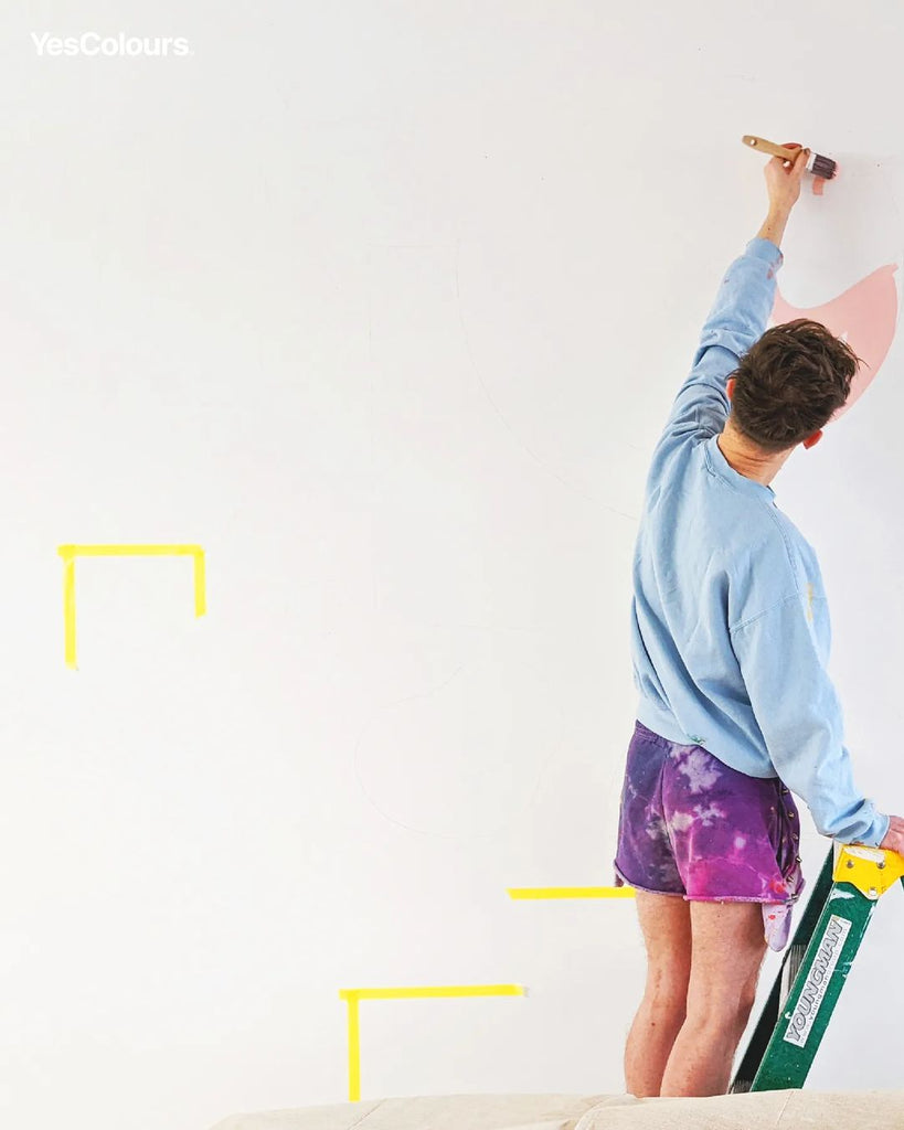 photo of a man climbed up on a step ladder and painting a white wall with colourful paint