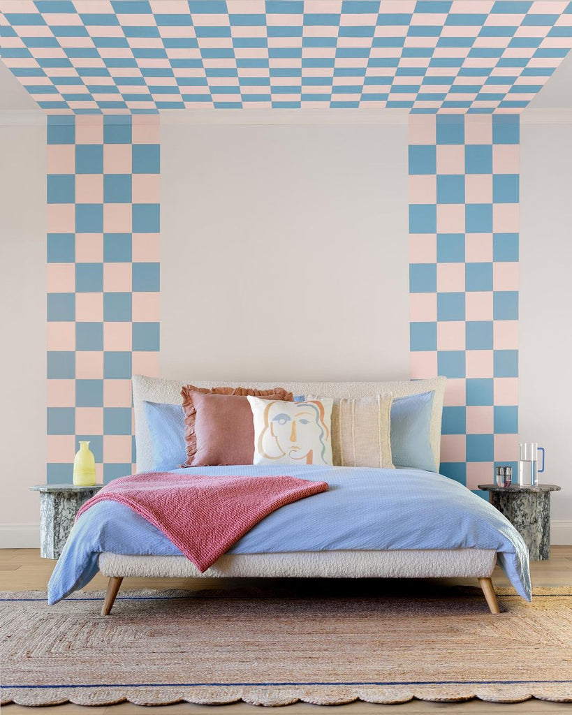 king size bed with blue bedding and a red throw, decorated with pillows and photographed in front of a checkerboard painted wall in blue, peach and neutral colours