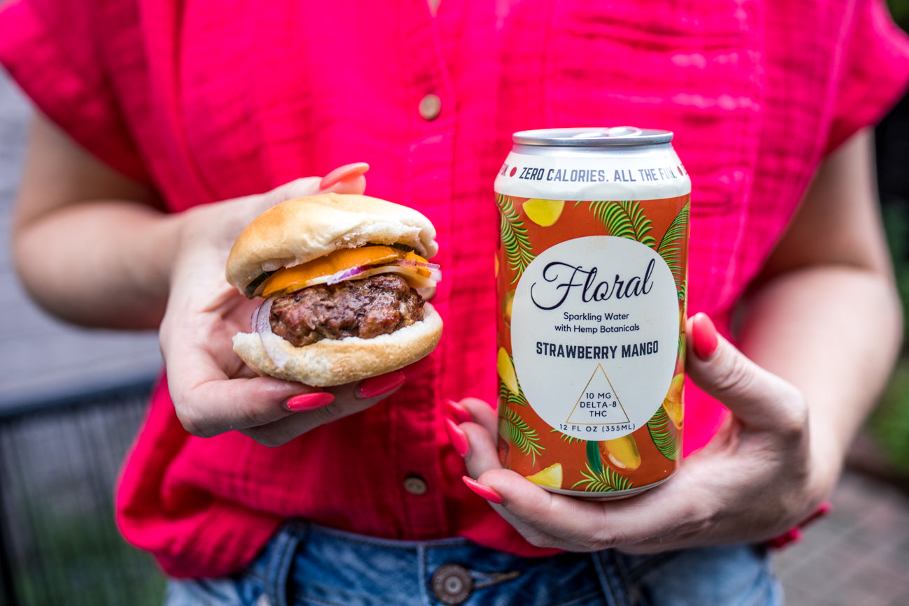 A person holding a hamburger in one hand and a can of Floral Beverages Strawberry Mango flavored THC Seltzer in the other hand.