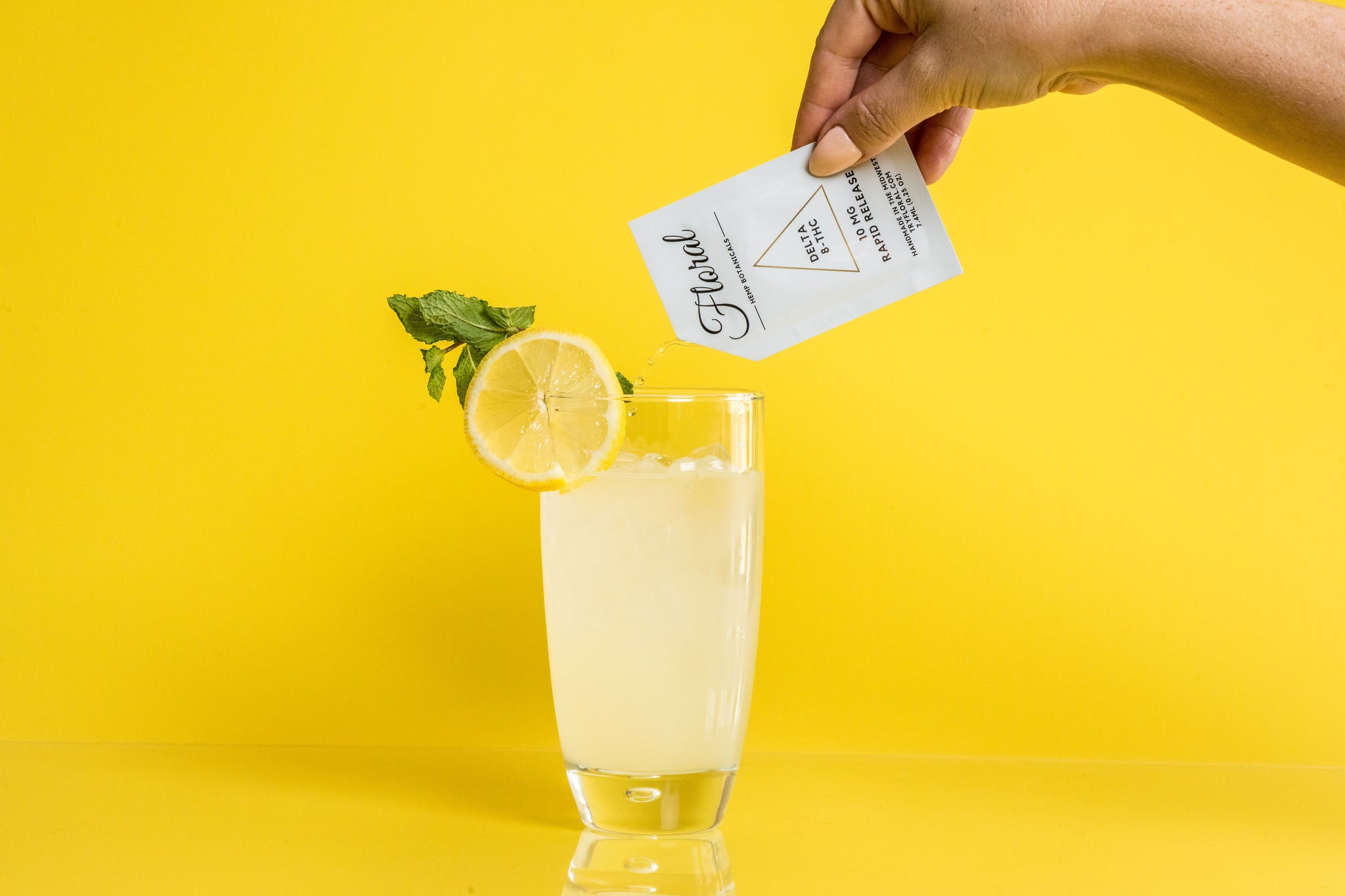 A hand pouring a packet of Floral Beverages' Floral Cocktail Enhancer into a glass of lemonade. The glass has a lemon wedge on it and is against a bright yellow background.