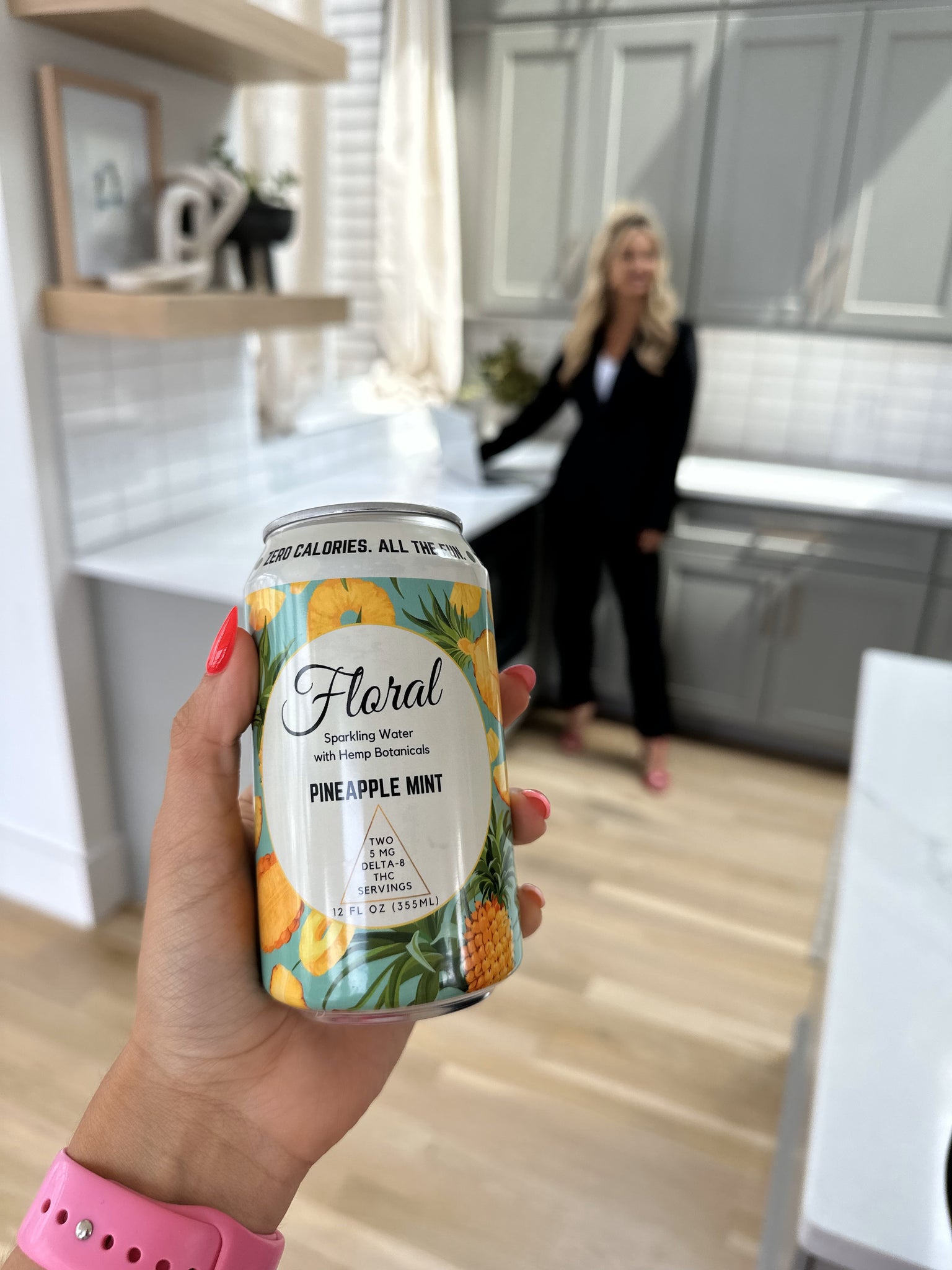 A women's hand holding a can of Floral Beverage's Pineapple Mint Sparkling Water in a kitchen with another woman in the background.