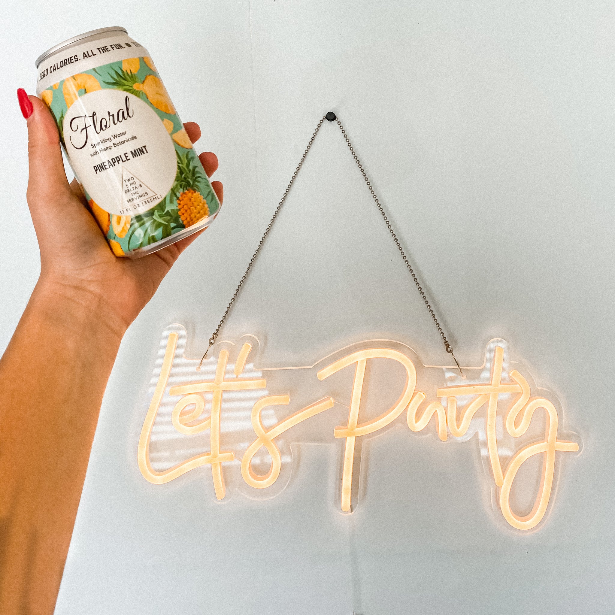 A hand holding a can of Floral Beverage's THC-infused sparkling water in the flavor Pineapple Mint in front of a neon-lit sign that reads "Let's Party."