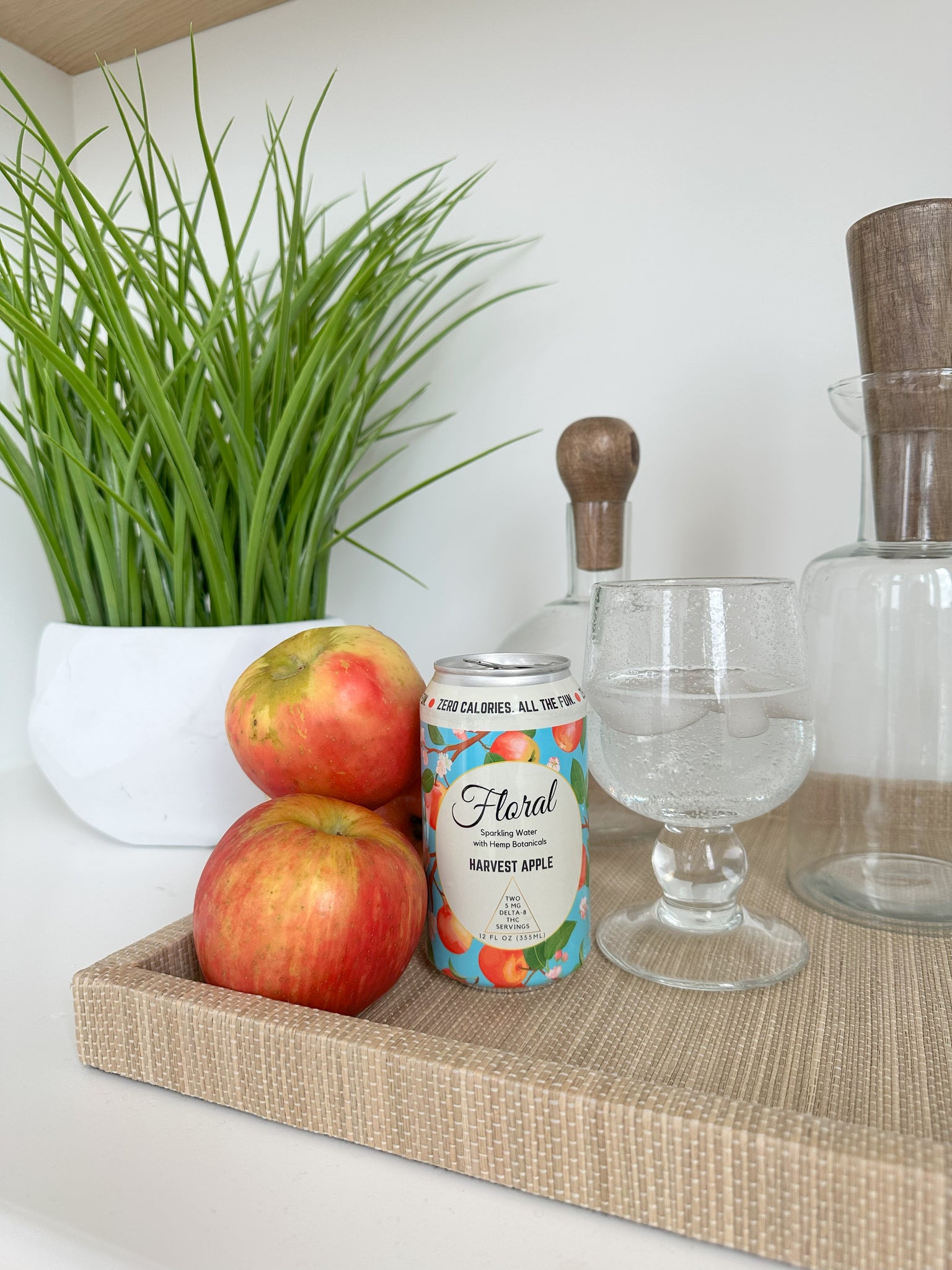 A can of Floral Beverages' Harvest Apple Sparkling Water sitting on a kitchen counter beside a cocktail glass and several apples.