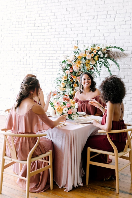 four women sitting around table in wicker chairs and velvet table cloth smile and cheers in revelry chiffon dresses