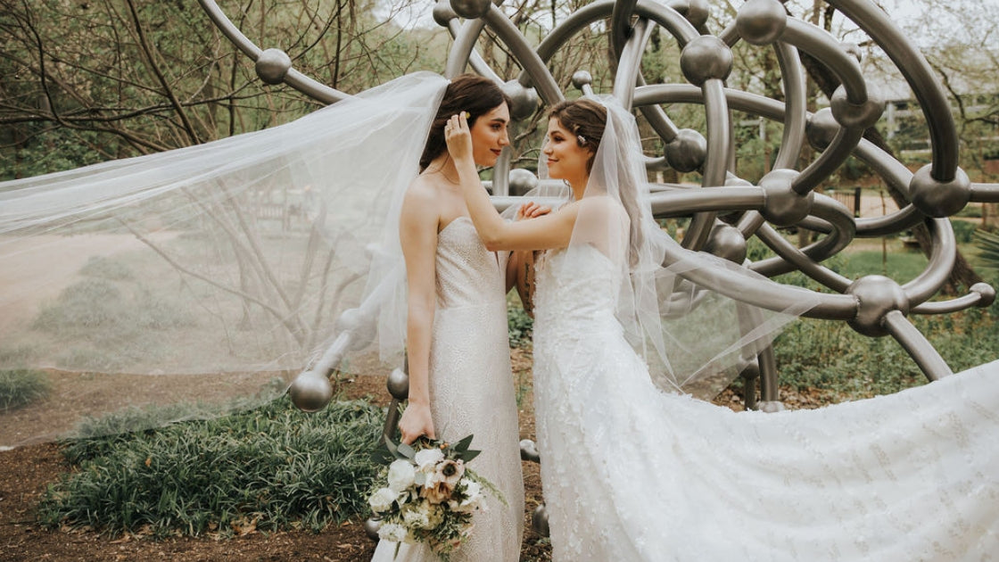 Two brides are better than 1 brides in revelry gowns veils wedding dresses holding flowers florals tucking hair behind ear