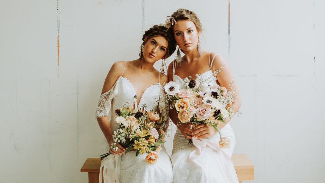 Two brides are better than 1 brides in wedding dresses wearing revelry bridal gowns sitting holding bouquets on wedding day