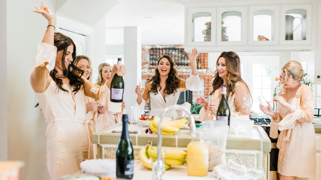 bride and bridesmaids getting ready on wedding day waring robes cheersing and smiling drinking champagne love