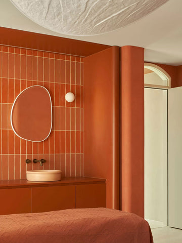 Tubb in Pastel Peach set against a backdrop of orange handmade Spanish subway tiles, complemented by Sussex Taps tapware in brass, custom orange curved cabinetry, and an organic-shaped mirror, creating a warm and inviting bathroom ambiance.
