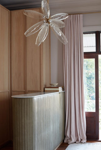 In the bedroom, a cozy ambiance is created through the use of timber cabinetry, adding warmth and character to the space. Pink drapes frame the windows, infusing a soft and romantic touch into the room. A glass light pendant hangs gracefully from the ceiling, casting a gentle glow and adding a touch of contemporary elegance to the bedroom's design.