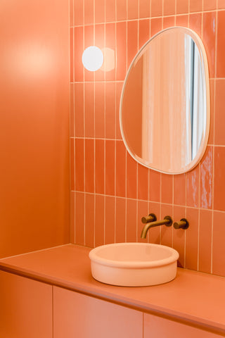 Another perspective of the bathroom, highlighting the Pastel Peach Tubb and orange cabinetry, with the warm glow of a wall sconce turned on, casting a soft light that accentuates the handcrafted Spanish subway tiles and the organic contours of the mirror.