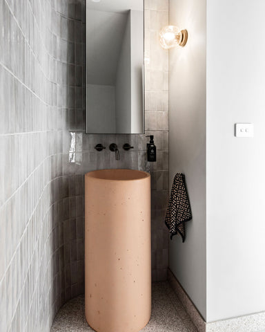 Luna Concrete pedestal basin in Pastel Peach sitting on grey and peach terrazzo tiles against a curved tiled wall with floor to ceiling Spanish Handmade Subway tiles in Grey with a turned on wall sconce and gunmetal tapware.