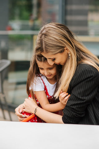 A color photograph of a mother holding her young daughter. The mother is standing on the left side of the image and is wearing a light blue shirt and jeans, while the daughter is standing on the right side of the image and is wearing a pink dress. The mother has her arms wrapped around the daughter, and the daughter is looking up at her with a big smile. The background consists of a green field and blue sky with some clouds. The image captures the love and bond between a mother and her child.