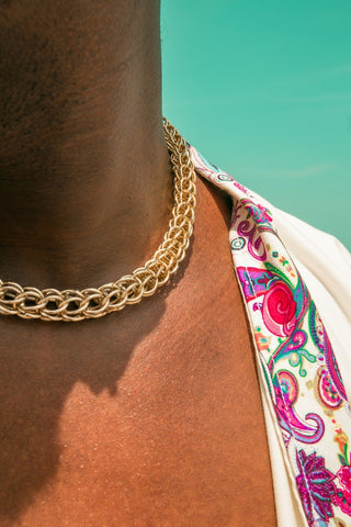 A close-up photograph of a person with dark skin wearing a silver chain choker necklace around their neck. The necklace is made of small interlocking links, and it is tightly fitted around the person's neck. The person is facing the camera, and their head and shoulders are visible in the image. The background is blurred, with no discernible details. The image captures the beauty and elegance of the necklace and the person's skin tone.