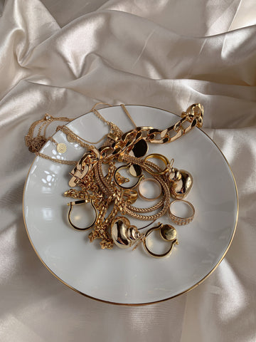 Assortment of gold jewelry including necklaces, bracelets, and rings arranged on a white plate, with their intricate designs and shimmering surfaces on display