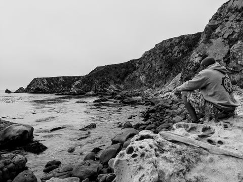 Black and White picture, rocky coastal cove, male sitting on shore holding a beer, wearing a hoodie with Buddhist symbol on back, camouflaged shorts, Cali8Folds