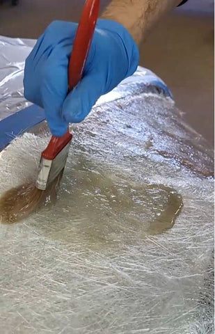 Gloved hand brushing polyester resin onto fiberglass chopped strand mat.