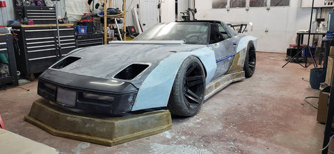 Custom corvette build with fiberglass and resin front and sides. Car has headlights removed and is sitting in garage surrounded by tools.