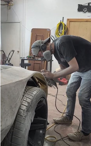 Man in respirator sanding fiberglass and resin side piece on car over tire.