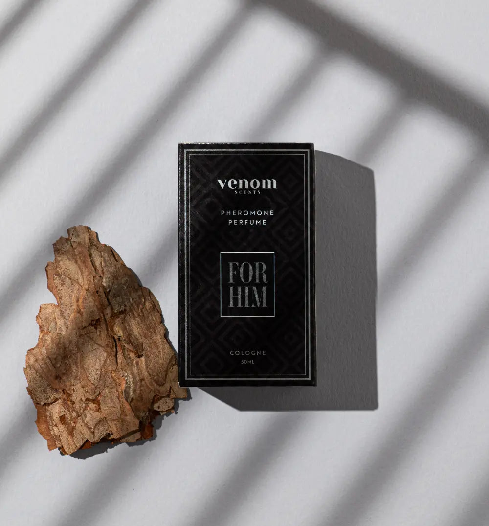 A perfume box labeled 'FOR HIM' next to a piece of bark, with shadow patterns on a light background.