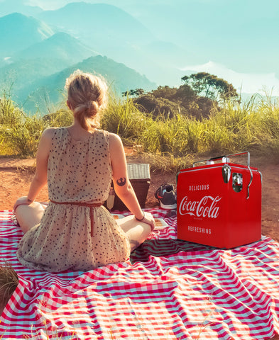 A woman wearing a dress and with a tattoo on her right arm, sits on a red and white checkered picnic blanket with only her back visible. Next to her is a red cooler ice chest, running shoes, a picnic basket, and some snacks. In the distance is green grass, mountains, and the sky.