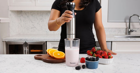 A person wearing a black t-shirt using the Kenmore stick blender to blend a light pink smoothie in the included blending jar
