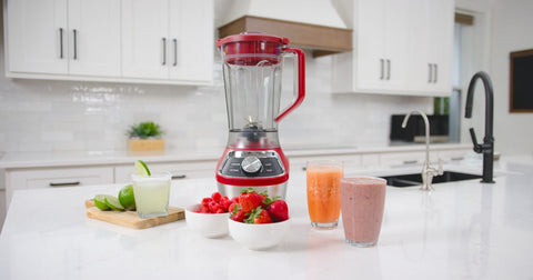 Kenmore stand blender, empty, on a white kitchen counter, with a wooden cutting board, glasses of smoothie and margarita, sliced limes, and bowls of berries arranged around it