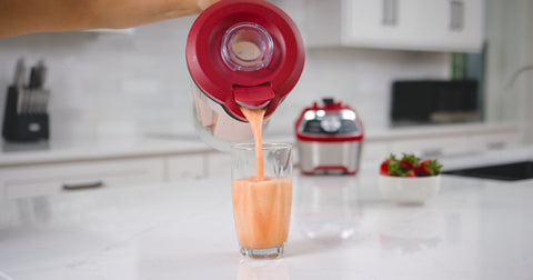 A person's hand holding up the red Kenmore countertop blender and pouring pale orange smoothie through the spout into a glass on a white kitchen counter