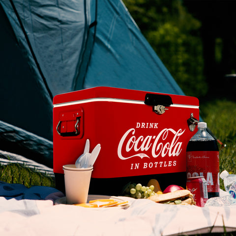 A red cooler with the words "Drink Coca-Cola in Bottles" written on it. The cooler is outside in front of a blue tent and it is sitting on a picnic blanket. Also on the blanket is a cup with utensils in it, grapes, an apple, a sandwich, and two bottles of cola.