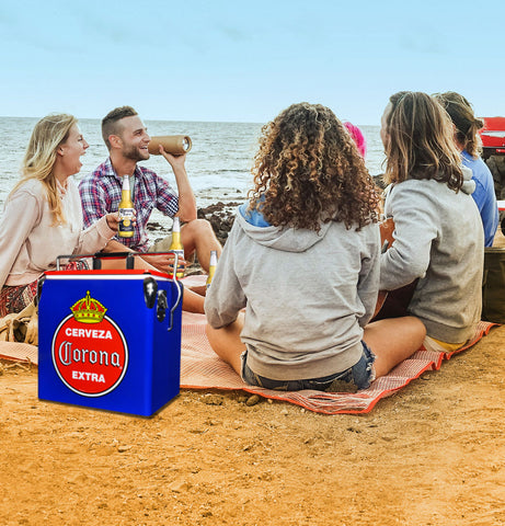 A group of people sit together on a blanket at a beach. There is sand under the blanket and water in the background. The people are holding beers and laughing. One man is holding a speaker. Next to the group of people is a blue and red ice chest box.