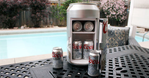 Photo of a can-shaped mini fridge, open and filled with cans of Coors Light beer, on a patio table with a swimming pool in the background