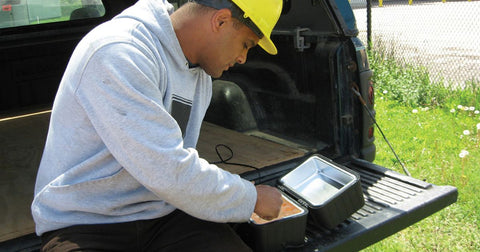 Photo of a man with medium brown skin wearing a gray hoodie and a yellow hardhat sitting on the tailgate of a truck eating from the lunchbox stove