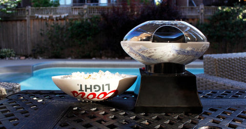 Lifestyle image of Coors Light popcorn maker with serving bowl filled with popcorn on a black patio table with a pool in the background