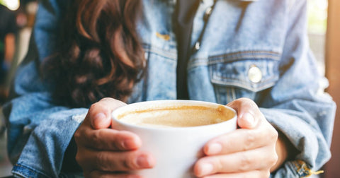 Closeup photo of a person wearing a blue shirt holding a white much full of coffee in their hands