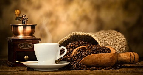 Photo of an old-fashioned hand-cranked coffee grinder, a white coffee cup, and an open burlap bag of coffee beans with a wooden scoop on a wood table