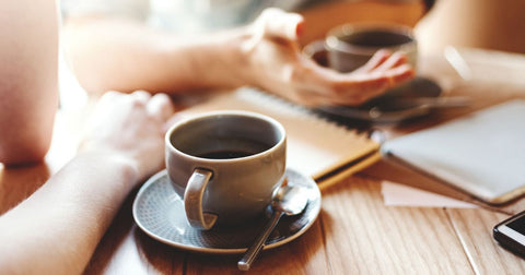 Closeup photo of two cups of coffee and a notebook on a wooden table. Two people's arms are visible, showing that they are sitting together drinking coffee and talking