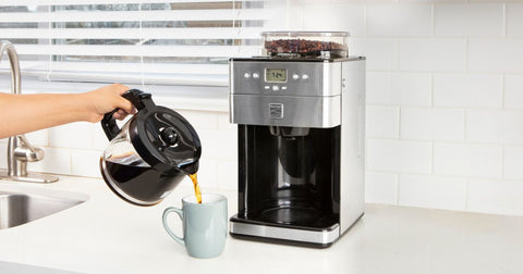 Photo of a person's arm pouring coffee from a glass carafe into a light blue mug in front of the Kenmore Grind and Brew coffee maker on a white kitchen counter