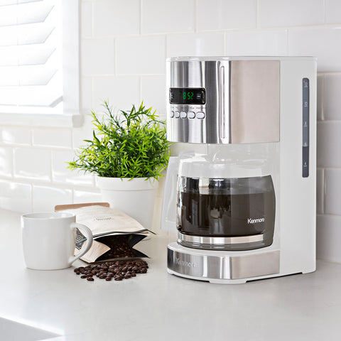 A white coffee maker with a full coffee pot sits on a white countertop. Next to the coffee maker is a green plant in a white pot, a bag of coffee beans spilled over revealing the beans, and a white coffee mug.