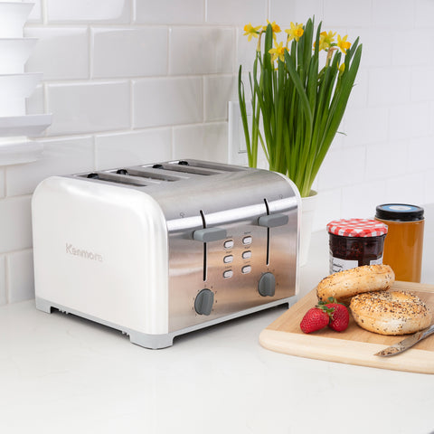 A white stainless steal 4-slice toaster sits on a white countertop. Beside the toaster is a green plant with yellow flower buds in a white pot, two jam jars, and two bagels, a knife, and strawberries on a cutting board.