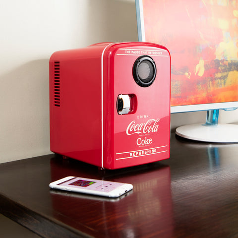 Dark red mini fridge with a black built-in speaker sits on a brown wooden desk with a smartphone and desktop computer beside it