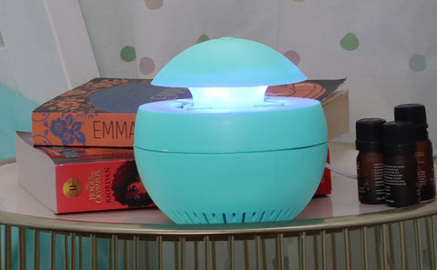 A blue mosquito trap sits on a table. Next to the mosquito trap are two books and three essential oils. Behind the table is a multicolored polkadot wall.