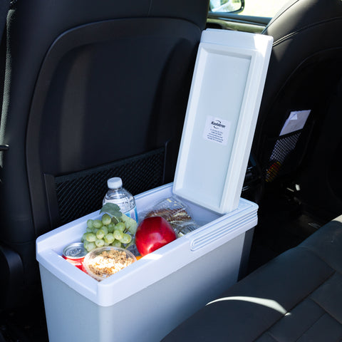 A white narrower cooler sits between the seats of a car. The lid is open and in the cooler are grapes, an apple, some granola, a pop can, and a water bottle.